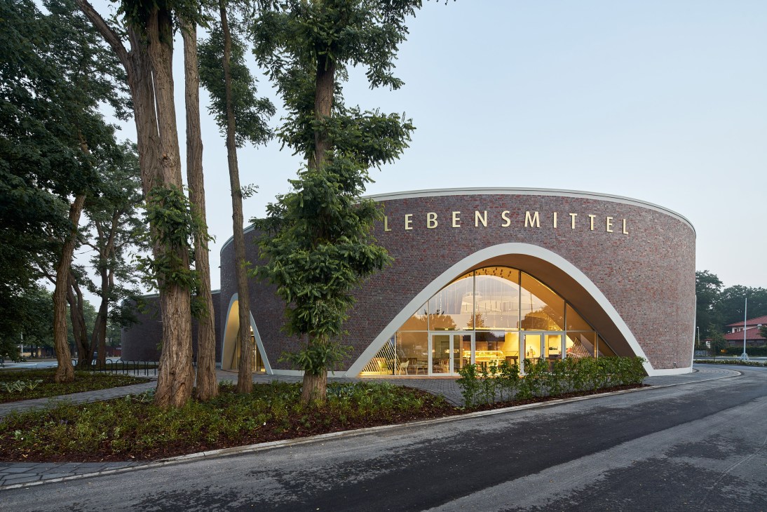 Modern red-brick supermarket building with curved white entrance and arched window, surrounded by tall trees in Oldenburg-Kreyenbrück, Germany.