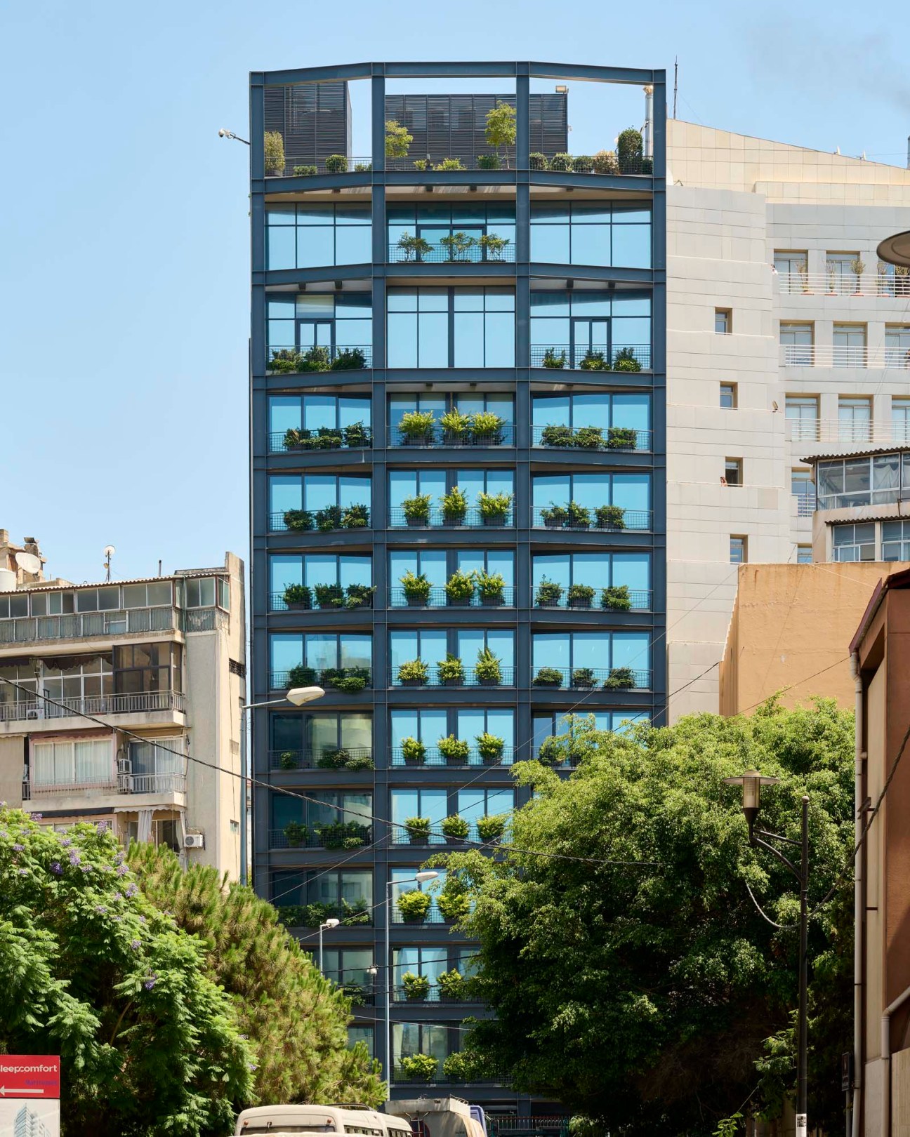 Modern residential tower with metal frame, glass windows, and plants on each balcony among older Beirut buildings