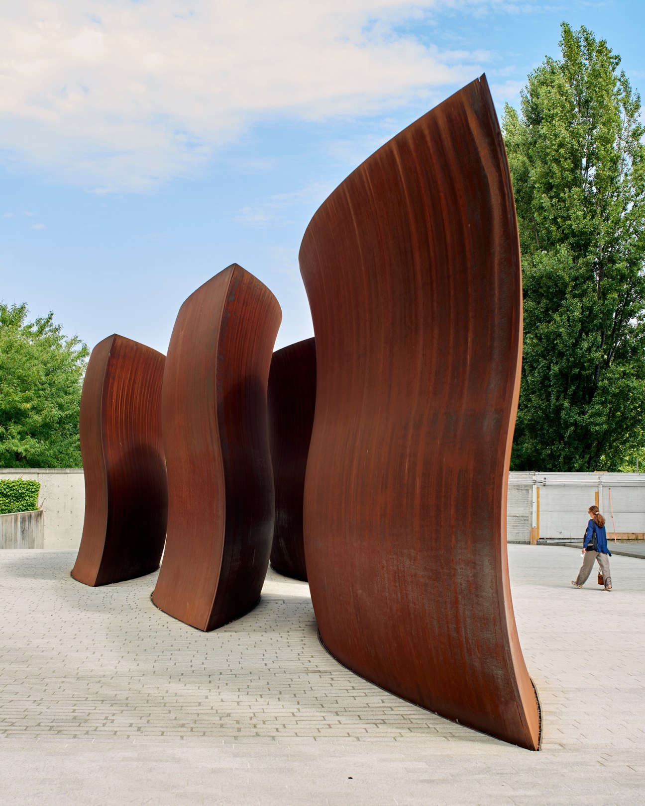 Large curved weathered steel sculpture by Richard Serra on paved courtyard with person walking nearby
