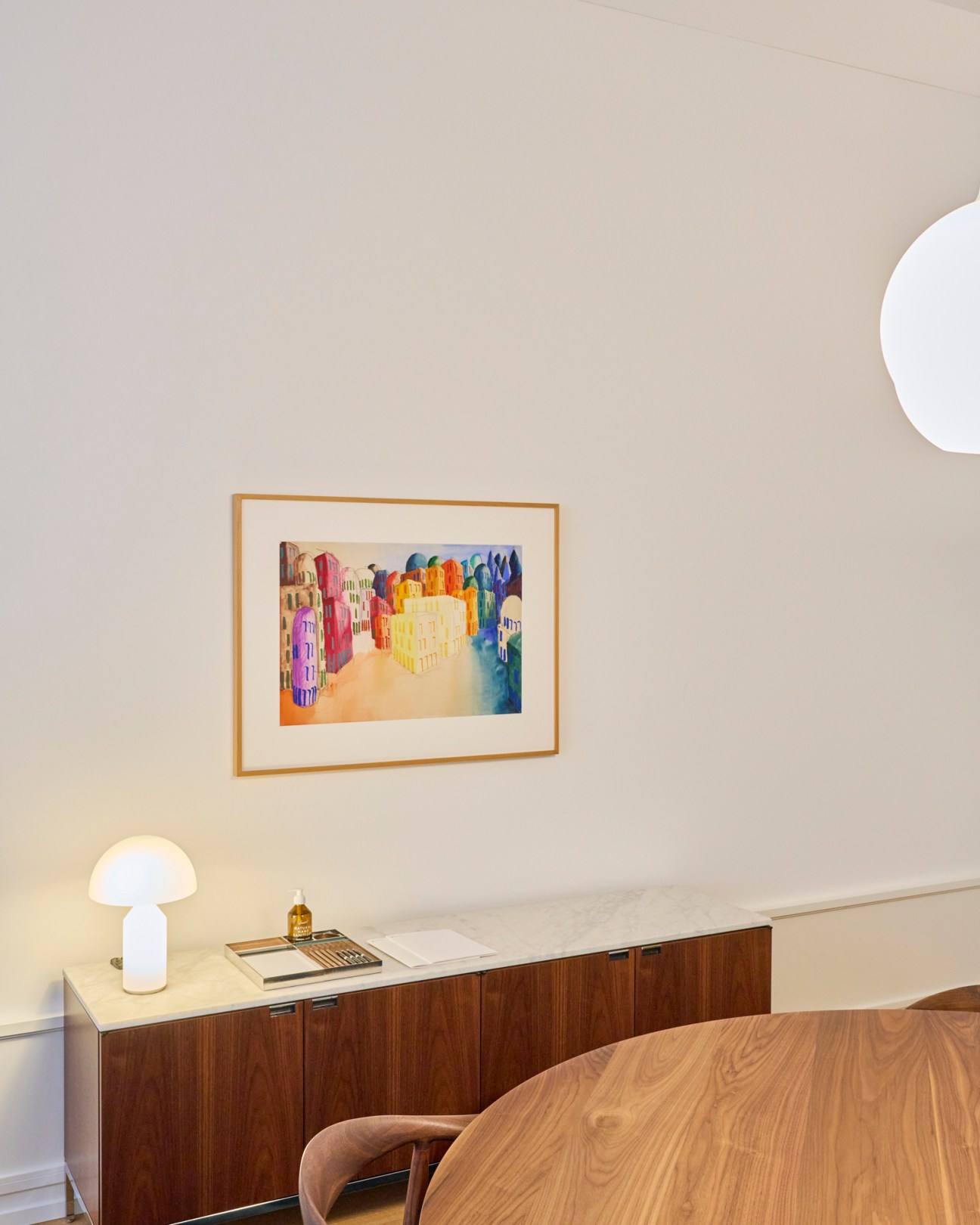 Framed colorful watercolor painting of urban buildings hanging above wooden sideboard with mushroom lamp