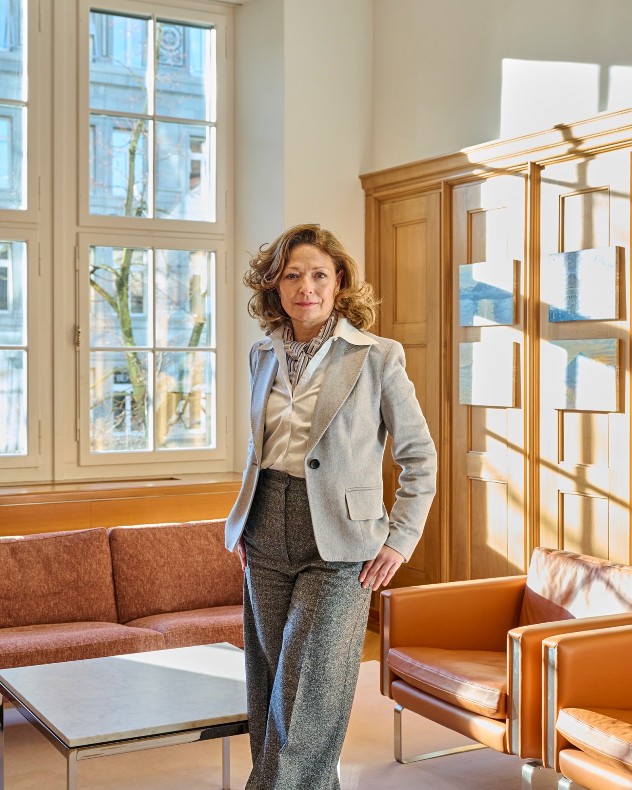 Woman in grey blazer and grey trousers standing in elegant office with tan leather furniture and large windows