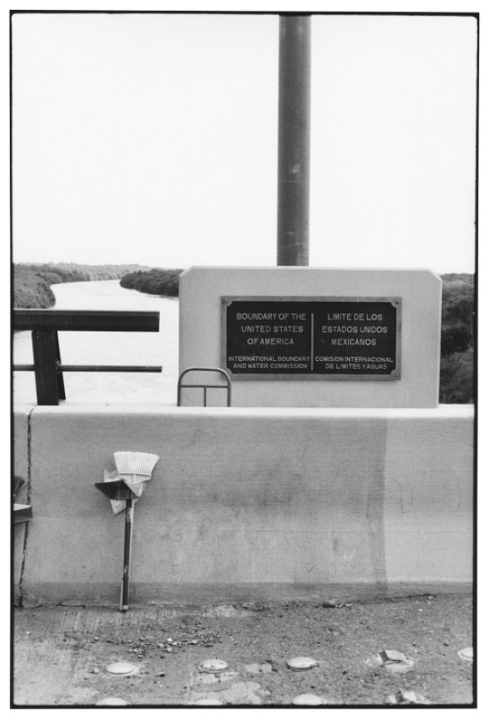 Black and white photo of US-Mexico border marker with bilingual plaque and broom leaning against concrete structure