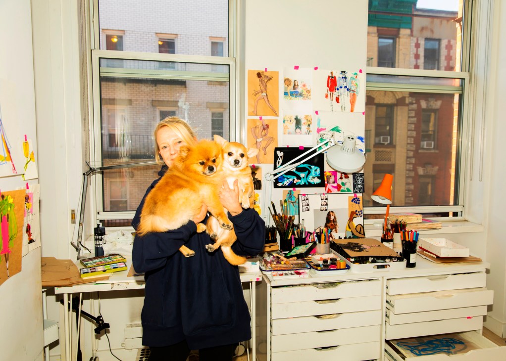 Jacky Marshall holding two small dogs in her New York studio workspace with art supplies and fashion sketches
