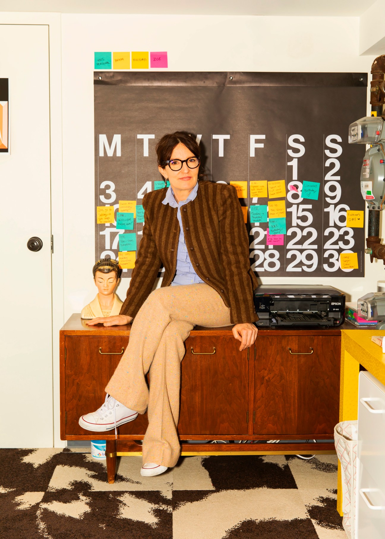 Set designer Kris Moran sits on a wooden desk in her Brooklyn home office, surrounded by a calendar with sticky notes and office supplies.