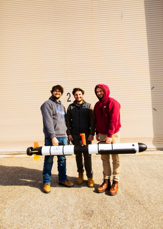 Three men holding a white torpedo-shaped drone outside a beige building