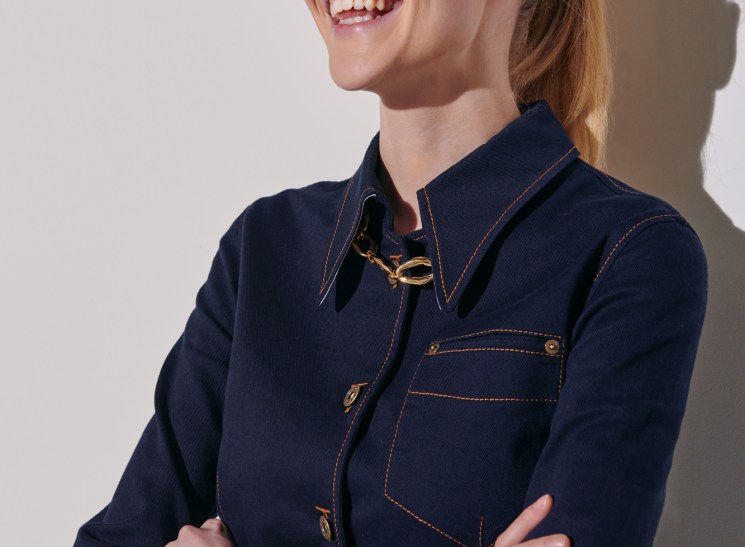 Woman in navy blue dress with gold jewelry and blonde hair smiling against white background