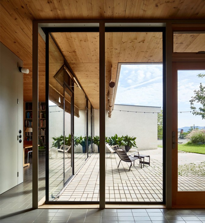 Open glass doors leading to a wooden terrace with lounge chairs and potted plants overlooking a landscape.