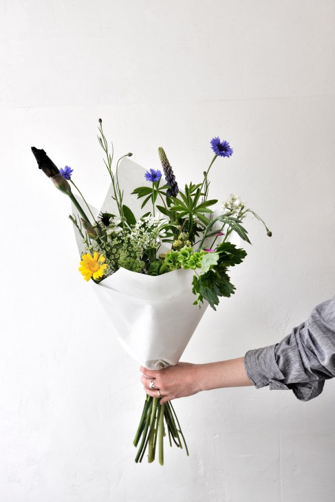 Hand holding a wildflower bouquet wrapped in white paper against neutral background