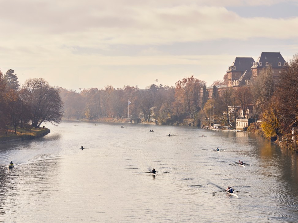 Rowers in boats on the River Po in Turin with historic buildings and autumn trees lining the riverbank.