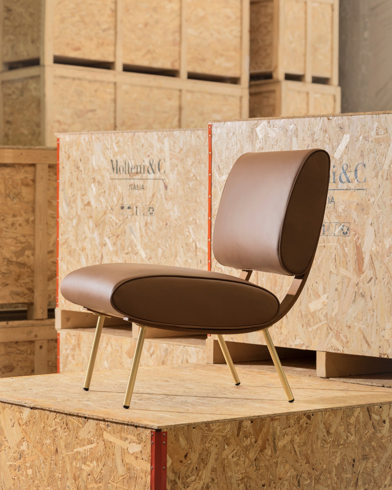 Brown leather chair with curved backrest and brass legs displayed on wooden platform in warehouse setting