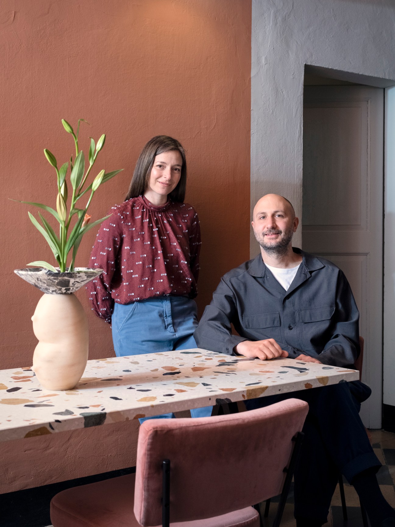 Two people posing in a minimalist interior with terracotta walls, a potted plant, and a textured table.