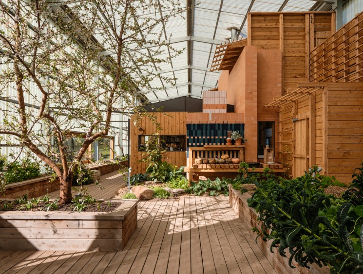 A sunlit indoor greenhouse with raised garden beds, mature trees, and a warm timber building structure beneath a glass roof.