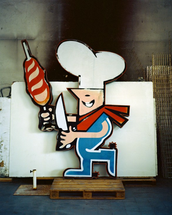 Cartoon chef character sign with white hat, blue apron, and red sausage for Chimarrão Brazilian steakhouse