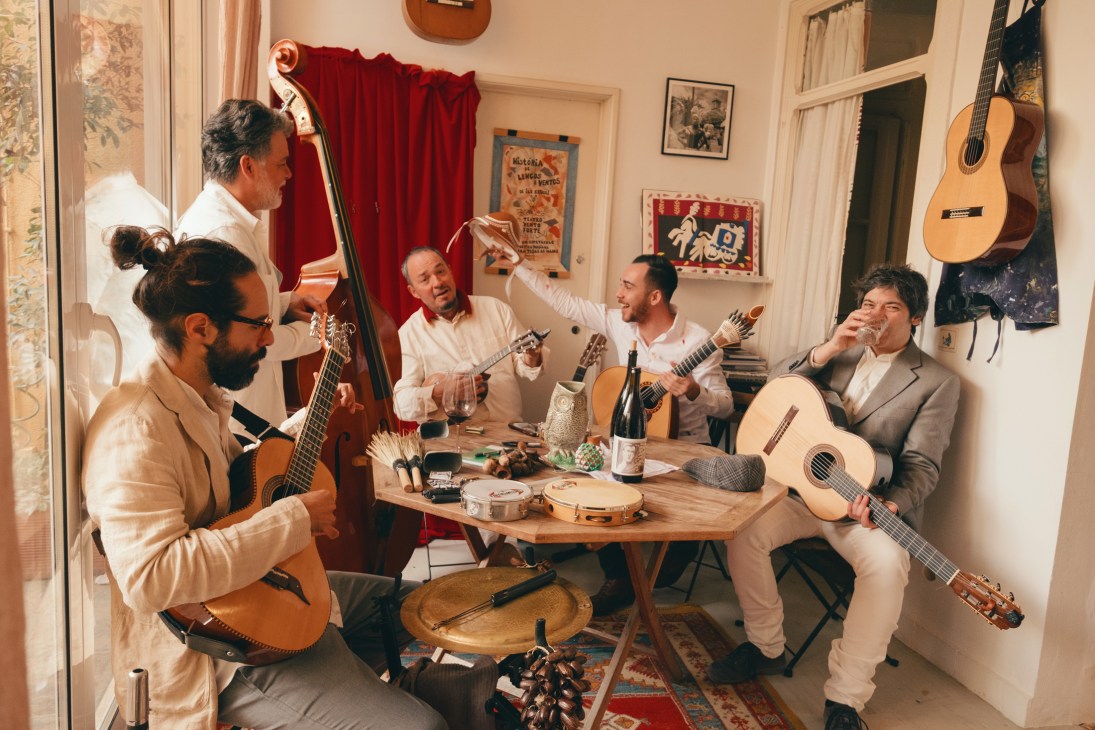 Five musicians with guitars and upright bass performing around a table in an intimate indoor setting