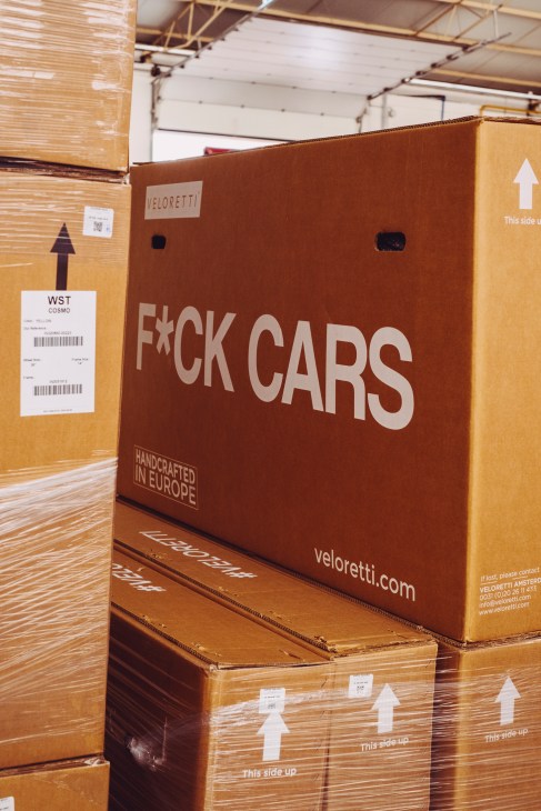 Large brown cardboard box with 'F*CK CARS' slogan and Veloretti branding in a warehouse with stacked packages