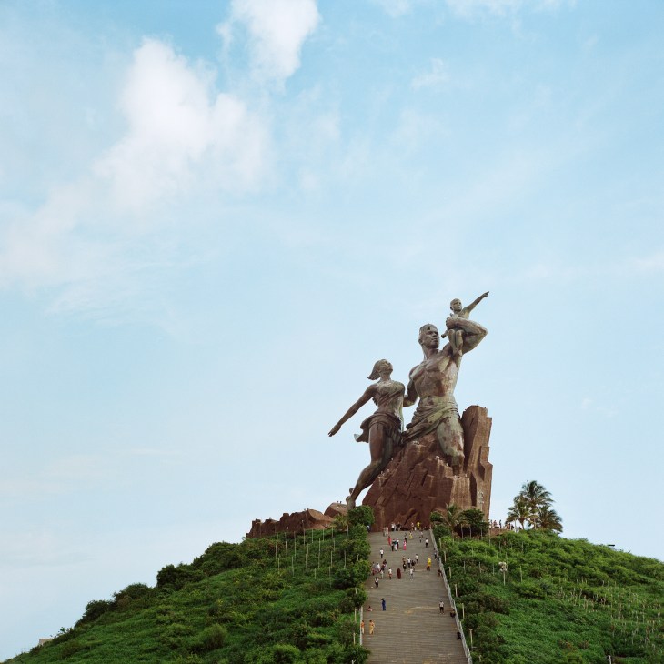 Large bronze monument depicting man, woman and child on hilltop with stairs and visitors in Dakar