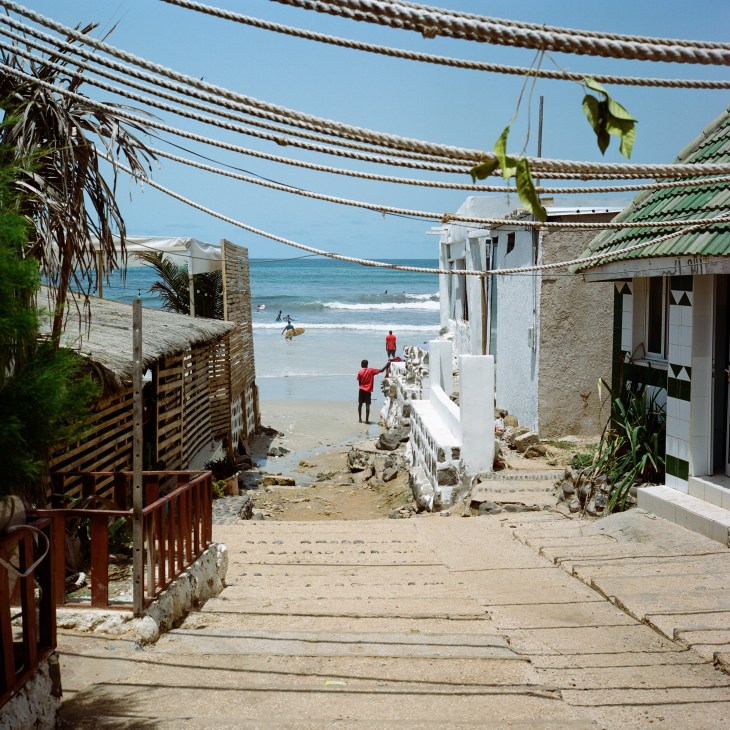 Stone steps leading down to a beach with ocean view, flanked by coastal buildings and tropical vegetation