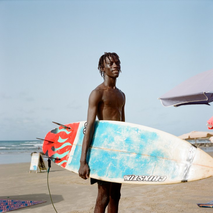 A shirtless young Black man with dreadlocks holds a surfboard on a beach