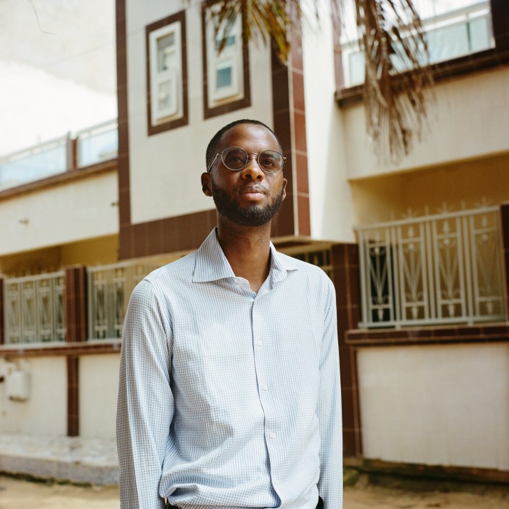Young Black man in glasses and light blue collared shirt standing outdoors in courtyard with colonial-style building