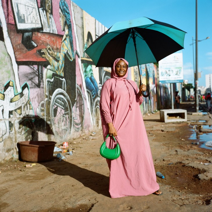 Woman in pink dress with headscarf holding green umbrella walking on street with colorful graffiti wall