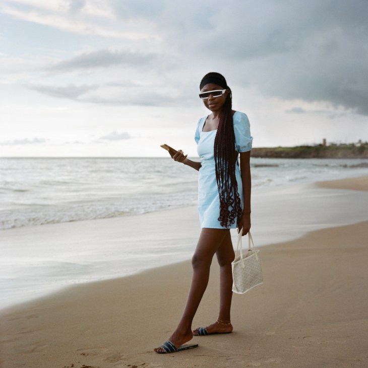 A woman in a light blue dress walks on a beach while looking at her phone