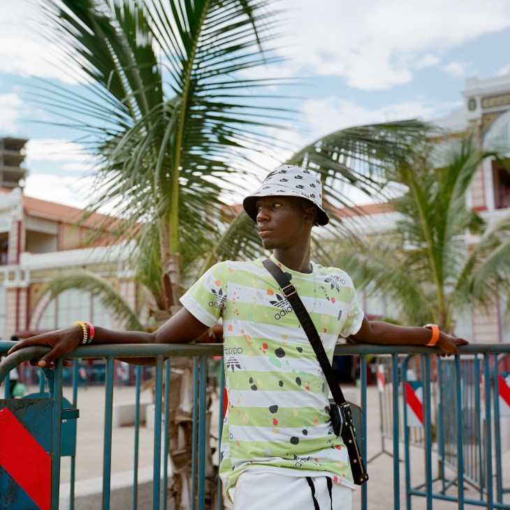 Young person in striped shirt and bucket hat standing by metal railings with palm trees in background