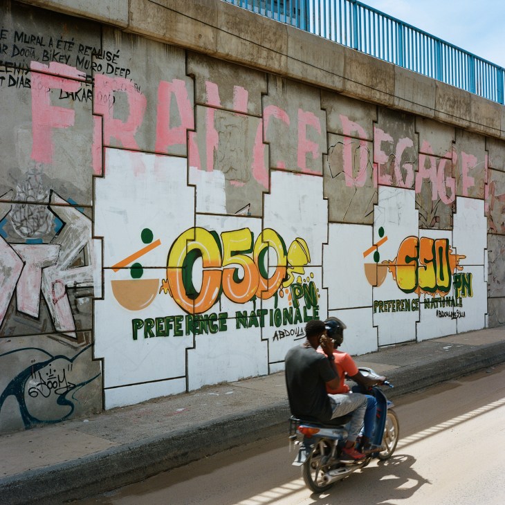 Anti-French graffiti on a concrete wall with two people on a motorcycle passing by