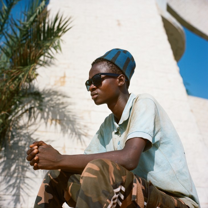 Young man in blue striped cap and sunglasses sitting outdoors with palm trees and white buildings in background