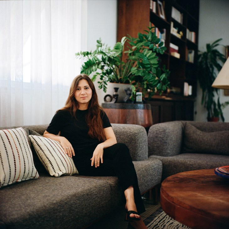 Océane Harati sits on a gray sofa in a modern living room with plants and bookshelves