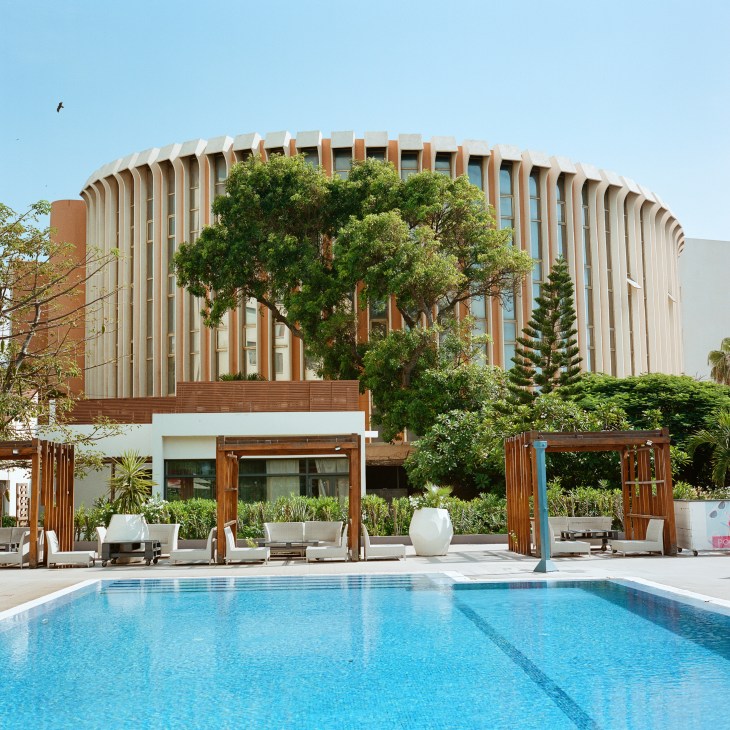 Modern curved building with vertical slats viewed from a swimming pool area with lounge chairs and umbrellas