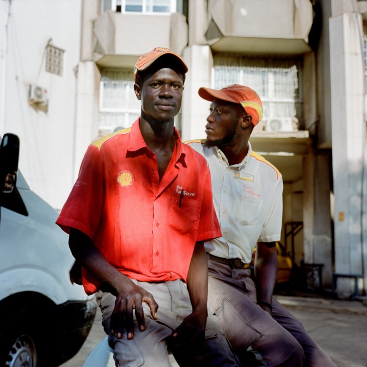 Two men in uniform sitting outside a gas station, wearing caps and work clothes