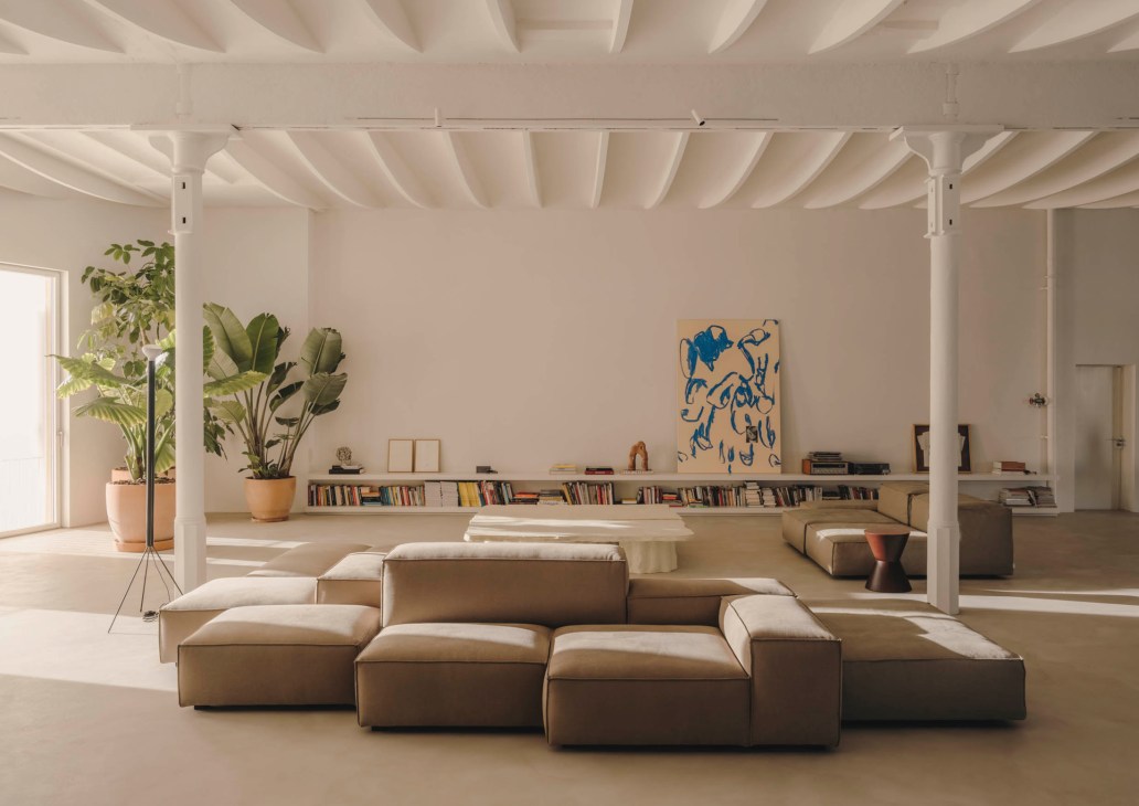 Modern interior with exposed white ceiling beams, modular tan seating, built-in bookshelf and large potted plants