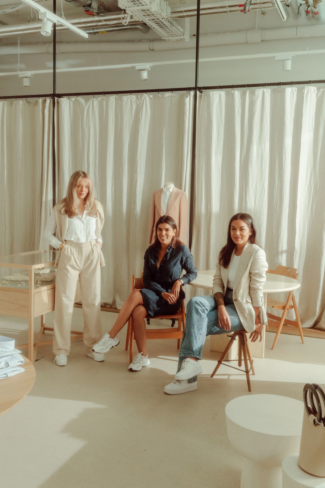 Three women in a light-filled tailoring studio with cream curtains and wooden chairs.