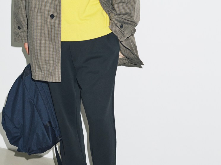 Model wearing grey checked coat over yellow jumper with navy trousers and Birkenstock sandals, carrying blue backpack