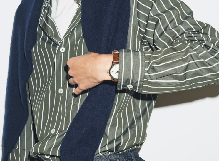 Model wearing navy jumper over striped olive shirt with brown leather watch, demonstrating autumn layering techniques