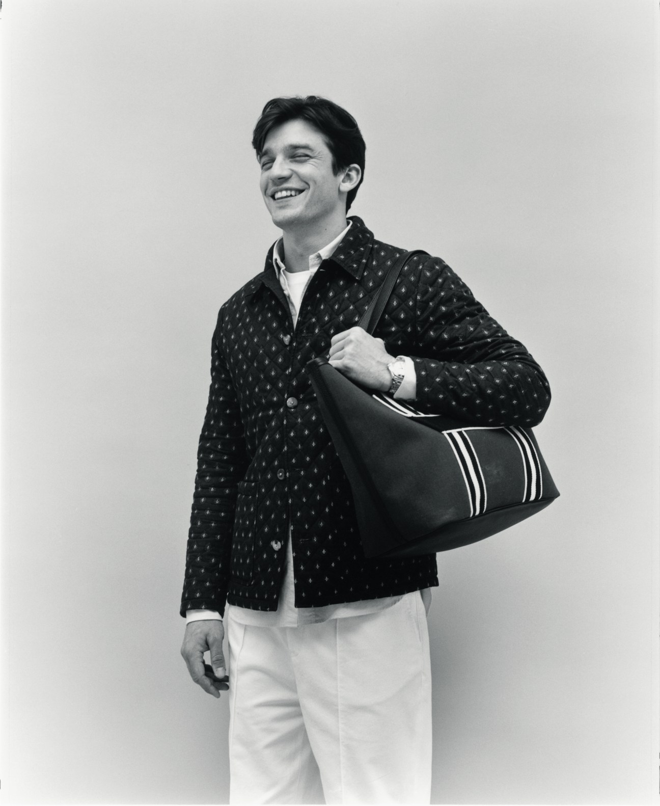 Man in patterned jacket and white trousers smiling while carrying a striped duffel bag