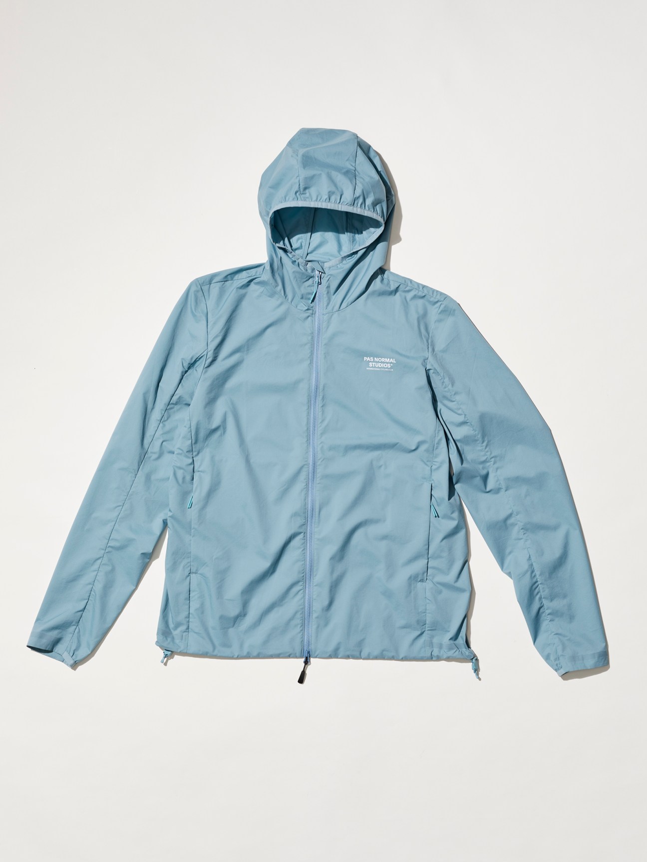 Light blue hooded cycling jacket with full zip closure and brand logo on chest which is a Pas  Normal Studios dusty blue ripstop cycling jacket with integrated storage pocket