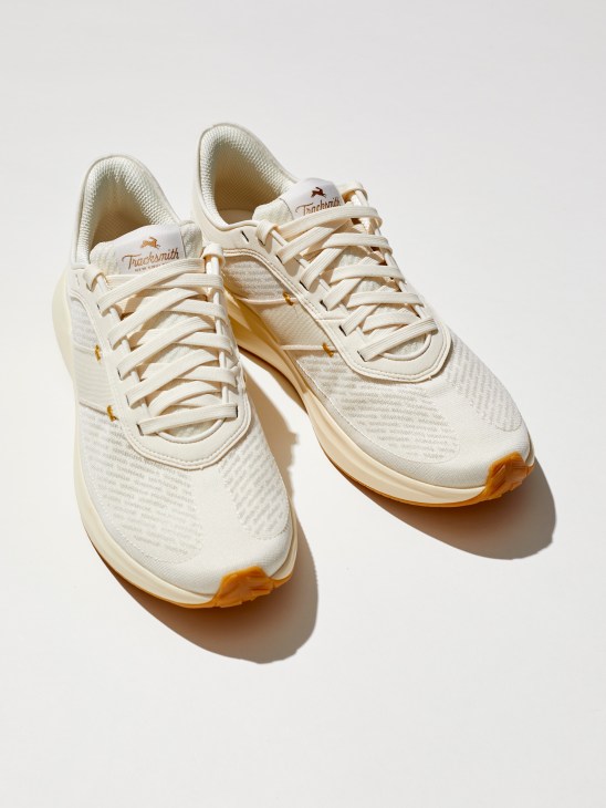 Pair of cream-colored Tracksmith Eliot Runner sneakers with woven mesh pattern and orange sole accents on white background