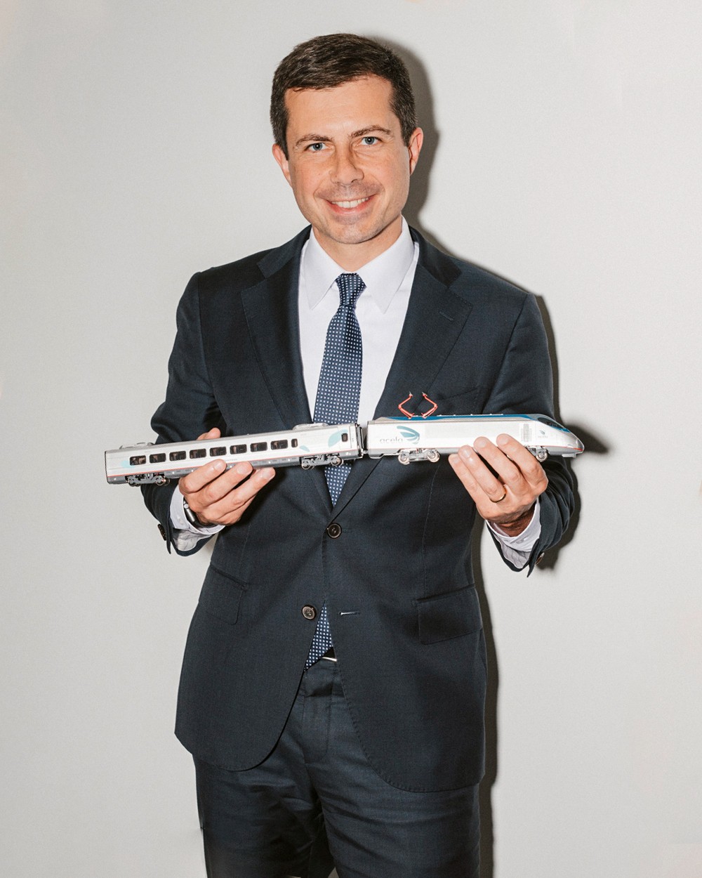 Pete Buttigieg in dark suit and blue tie holding a model passenger train against white background