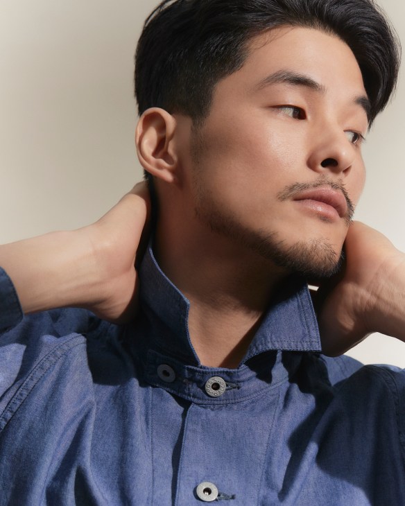Asian man in blue denim shirt with hands behind neck looking to the side in contemplative pose