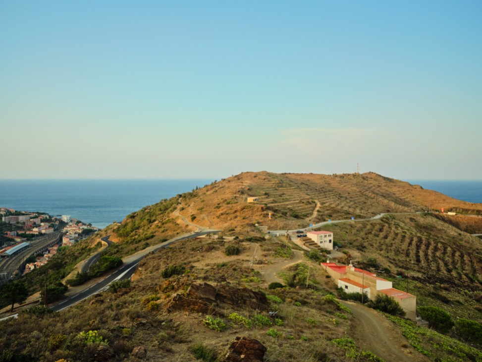 Aerial view of a winding road crossing a hillside between coastal towns, with the Mediterranean Sea in background