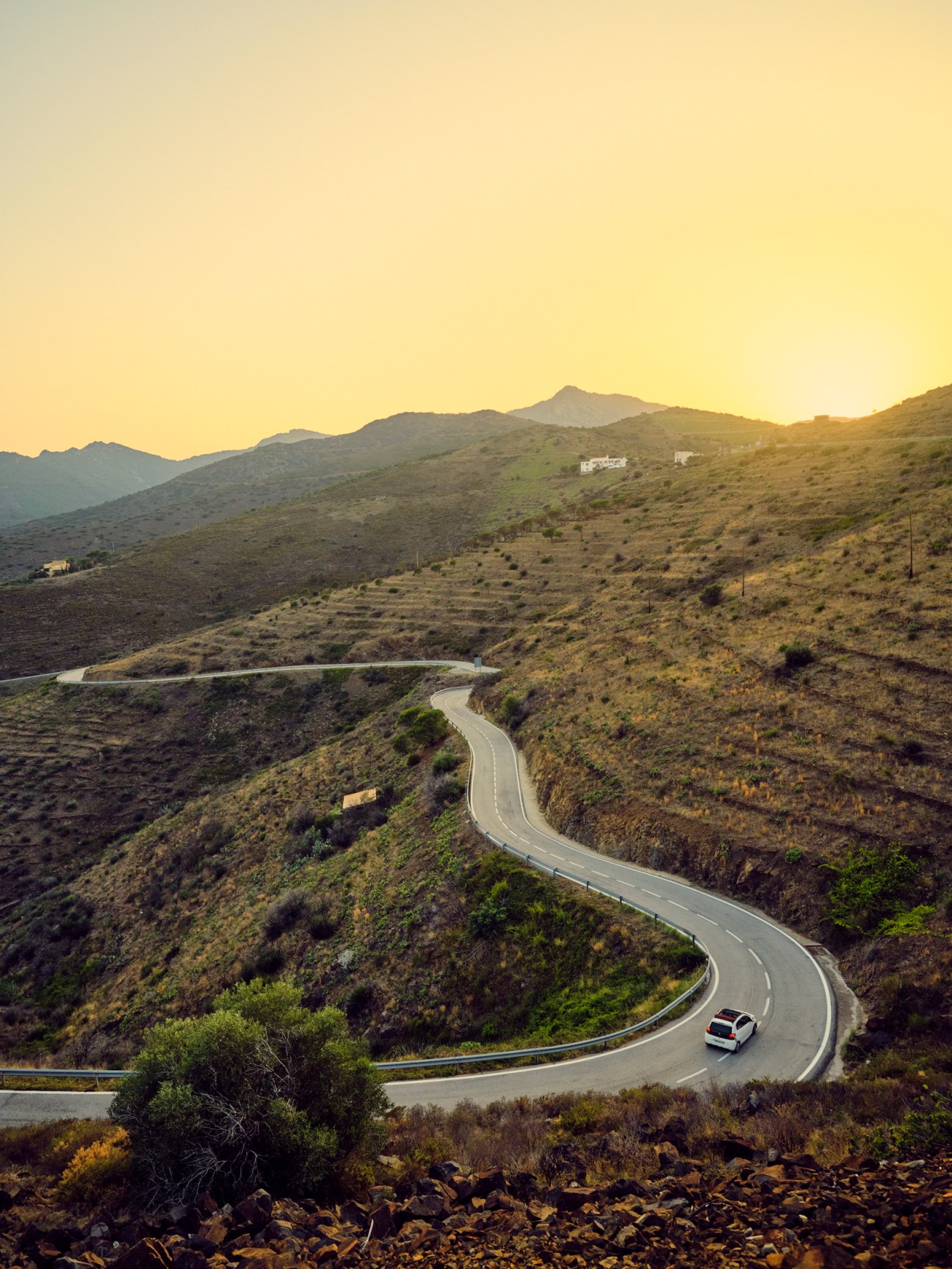 A winding mountain road curves through arid hills at sunset with a single car traveling the scenic route