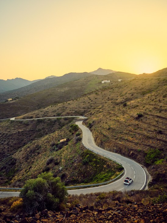 A winding mountain road curves through arid hills at sunset with a single car traveling the scenic route