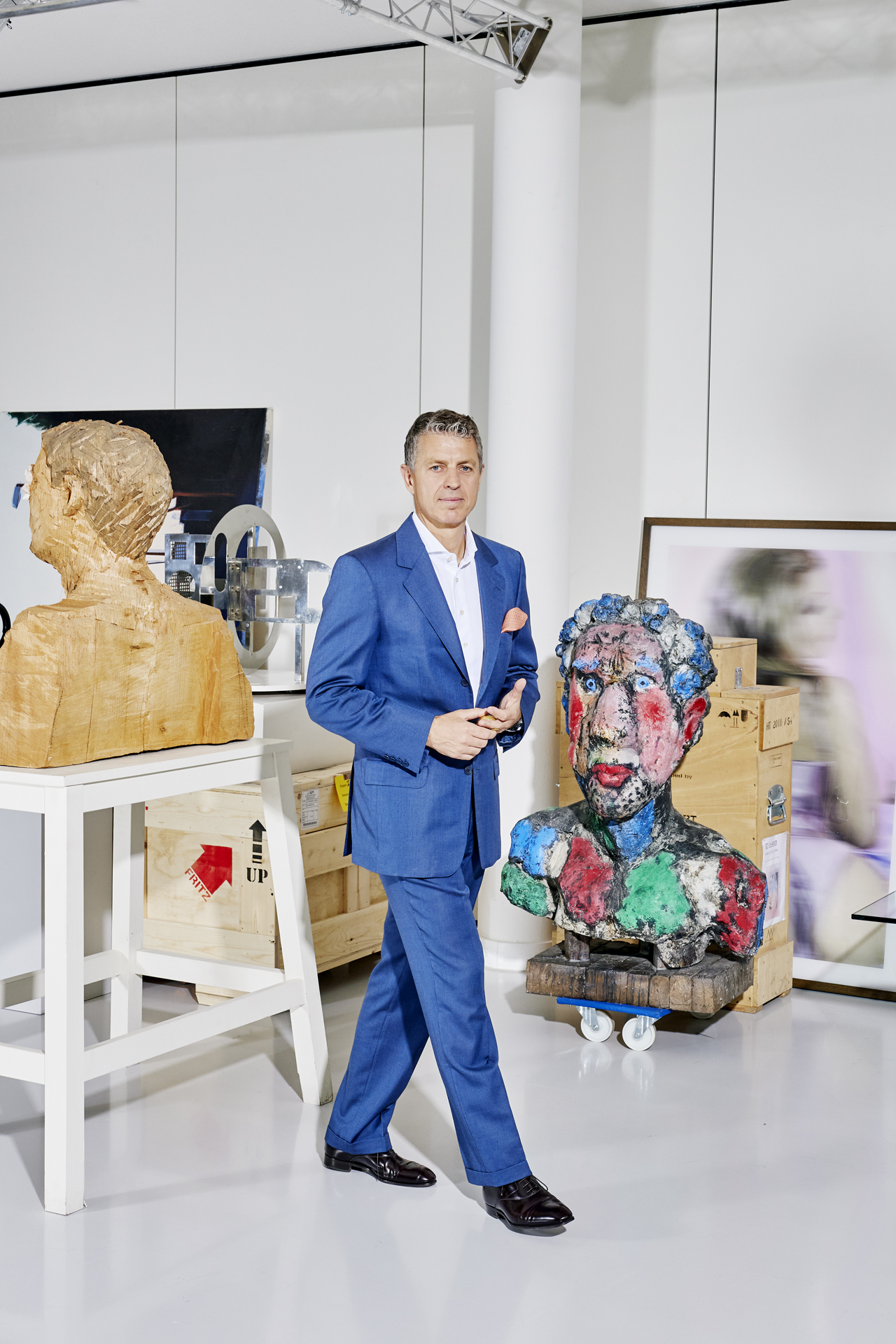Man in blue suit standing in gallery space surrounded by sculptural busts and artworks.