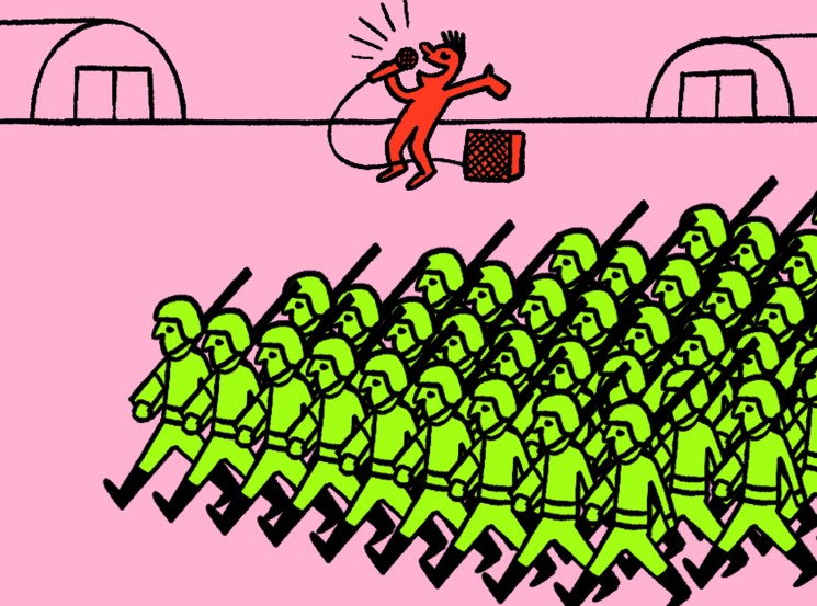Red figure with microphone performing on stage above rows of green uniformed soldiers with rifles in formation