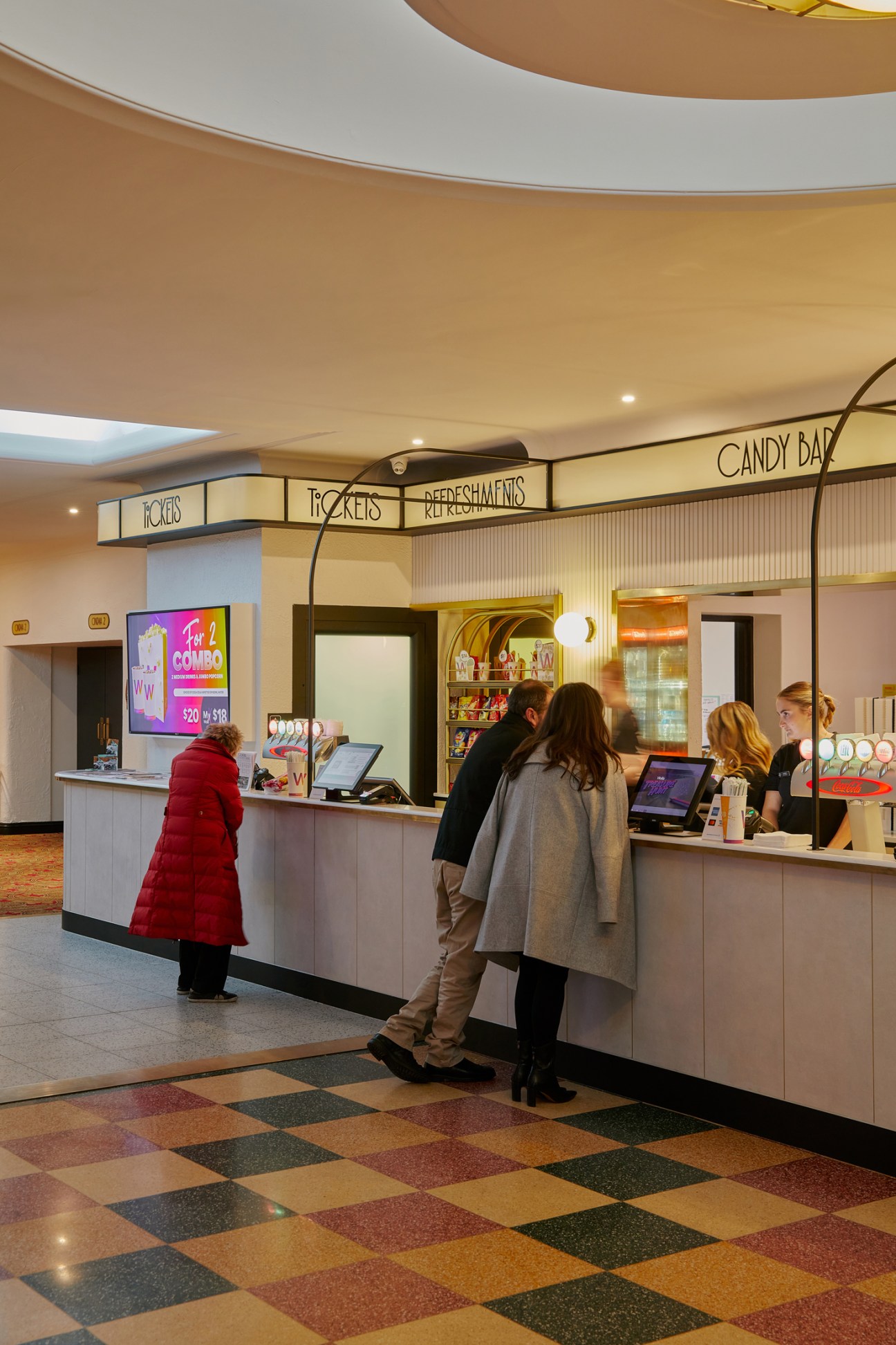 Cinema concession stand with curved counter, colorful checkered floor, and customers purchasing tickets and refreshments