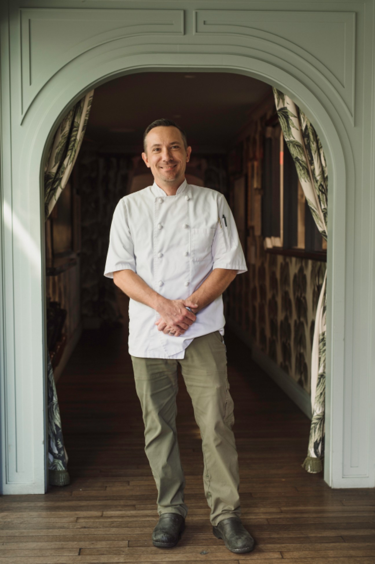 Chef Tim Morton in white chef's coat standing in arched doorway with hands clasped