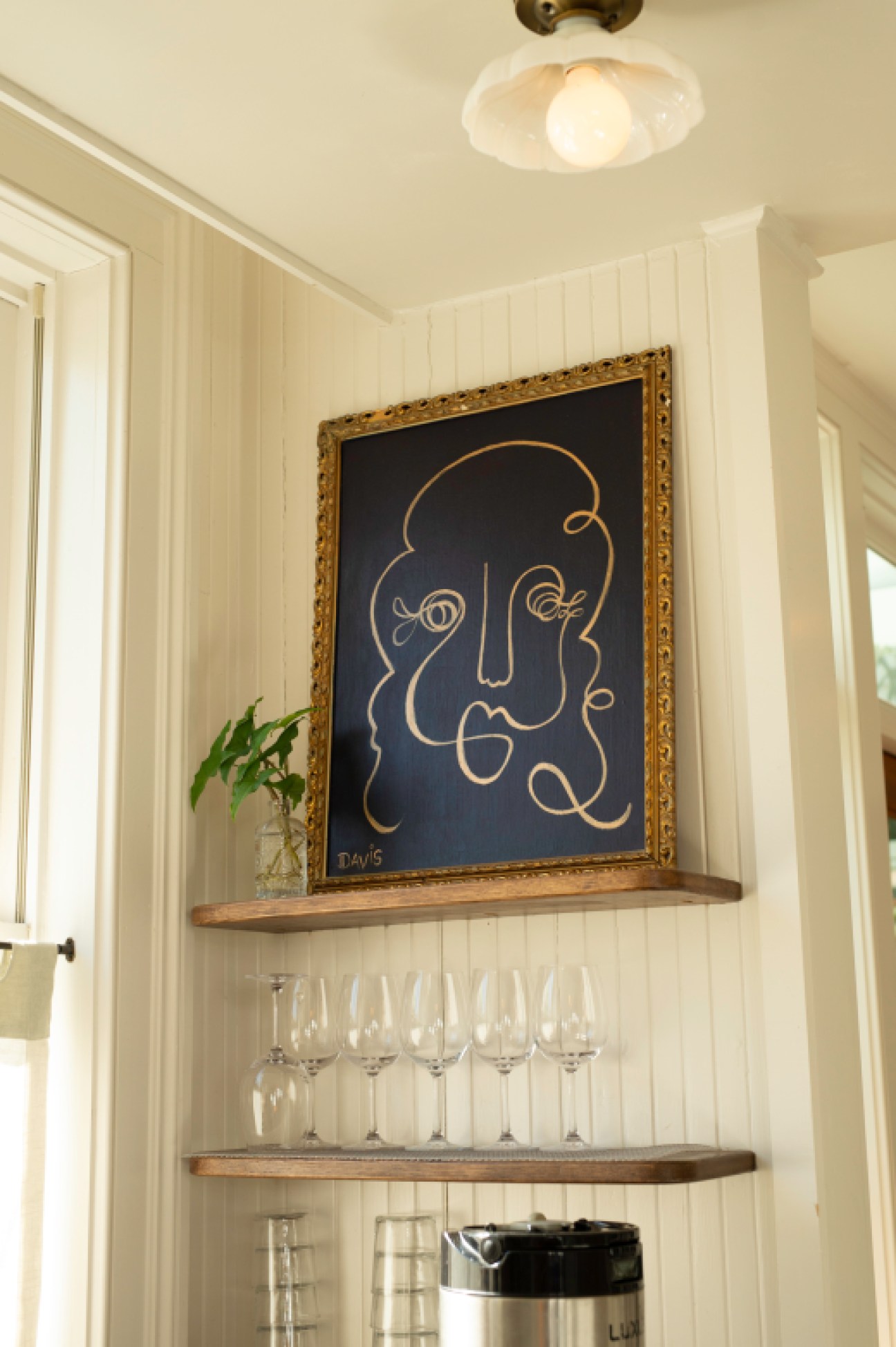 Ornate framed artwork depicting a face in gold lines on dark background above shelves with wine glasses