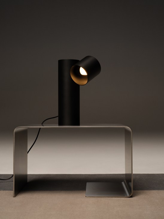 Black cylindrical table lamp with angled light head positioned on clear acrylic side table against gray background