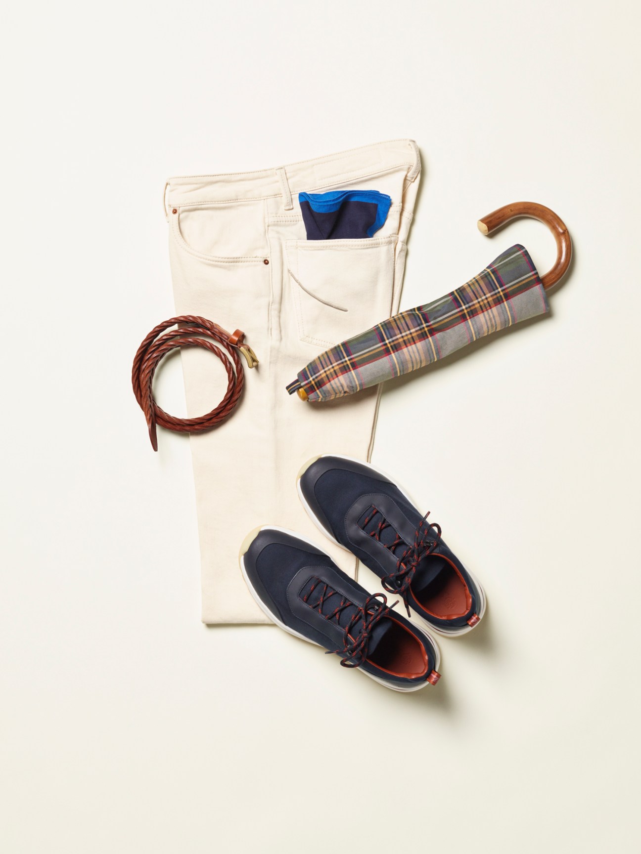 Flat lay of cream jeans, braided brown belt, plaid umbrella, and navy sneakers on white background